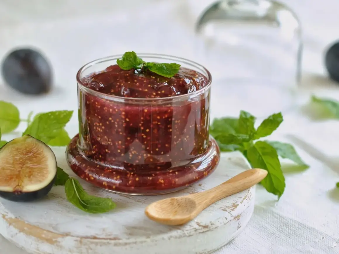 Tarro de mermelada de higos casera, con higos frescos y hojas de menta alrededor. Una cuchara de madera descansa cerca.