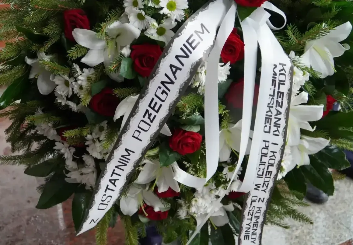 Wieniec z białych lilii, czerwonych róż i zieleni. Na białej szarfie widnieje oryginalny napis na szarfie pogrzebowej: "Z ostatnim pożegnaniem Koleżanki i Koledzy z Komendy Policji w Olsztynie".