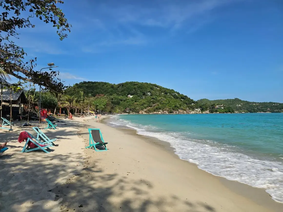 Solig strand på Koh Phangan, Thailand. Turkost vatten, gröna kullar och tomma solstolar väntar på besökare.
