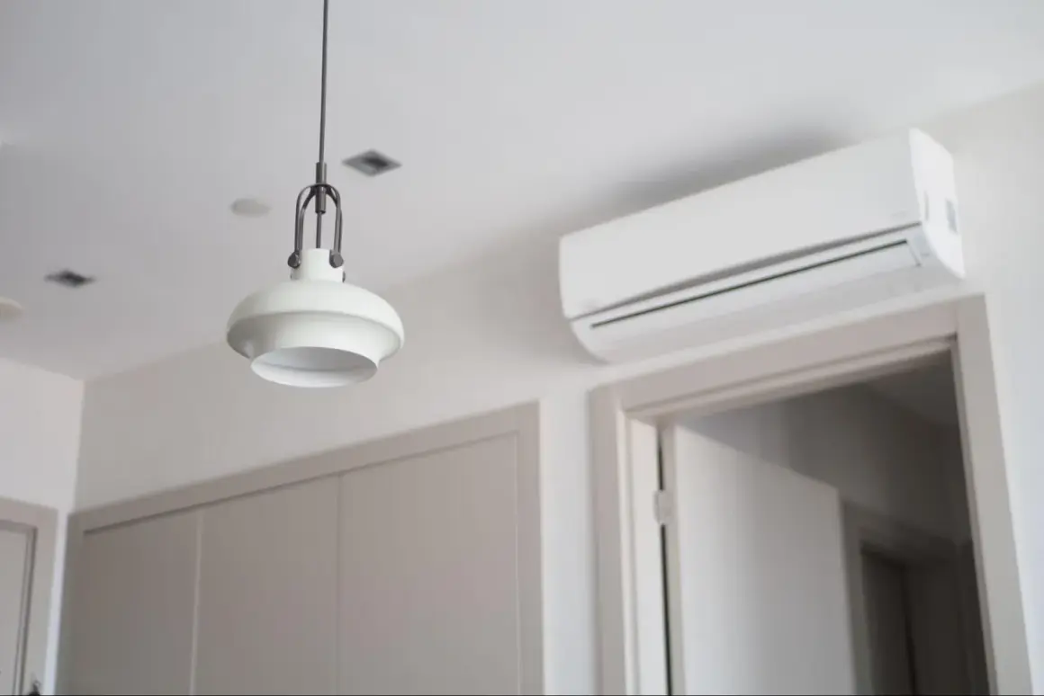 Biała jednostka klimatyzacyjna zamontowana na ścianie nad drzwiami, obok lampy wiszącej.