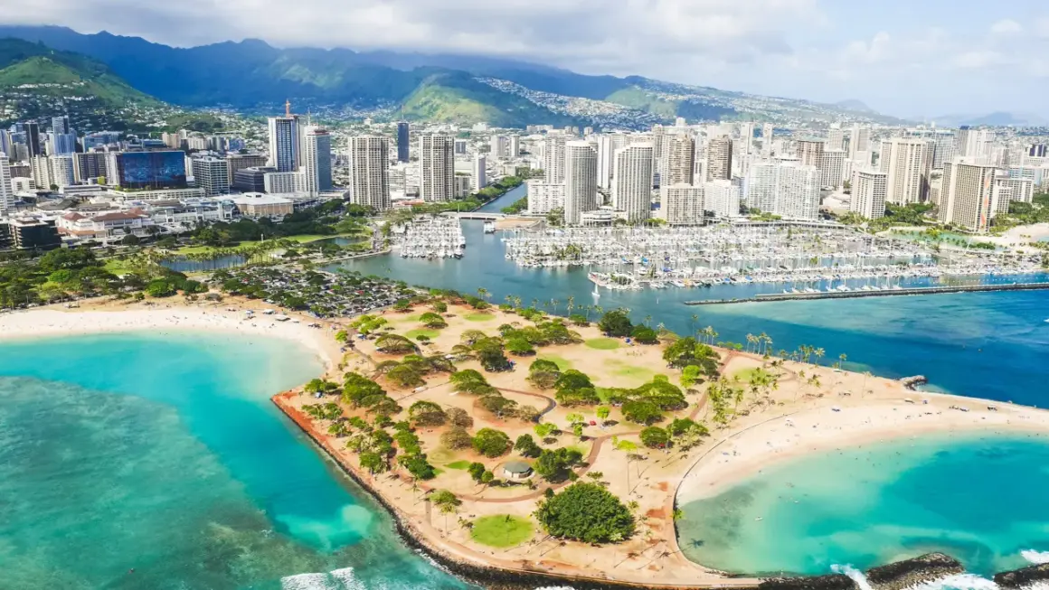 Widok na Honolulu, stolicę Hawajów, z plażą, parkiem i mariną pełną jachtów. W tle góry i drapacze chmur.