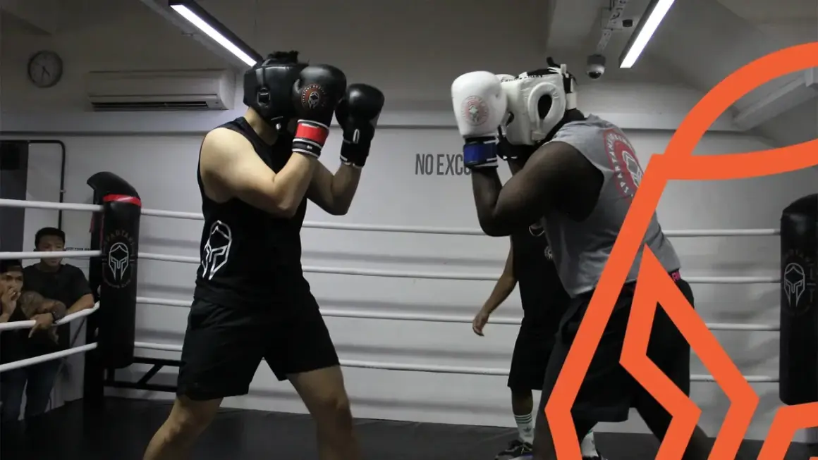 Two boxers spar in a ring, practicing their boxing defense. One boxer wears a black tank top and headgear, the other a grey tank top and white headgear.