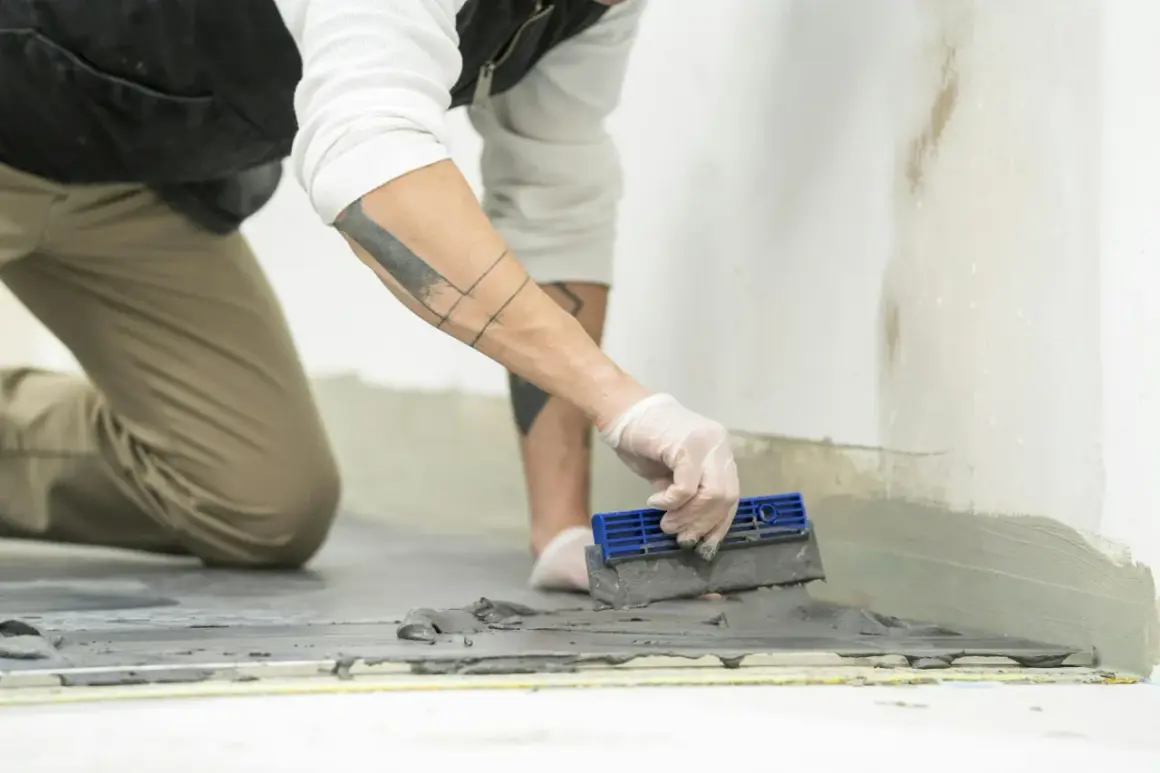 Un operaio con guanti bianchi sta stuccando piastrelle sul pavimento con una spatola blu.