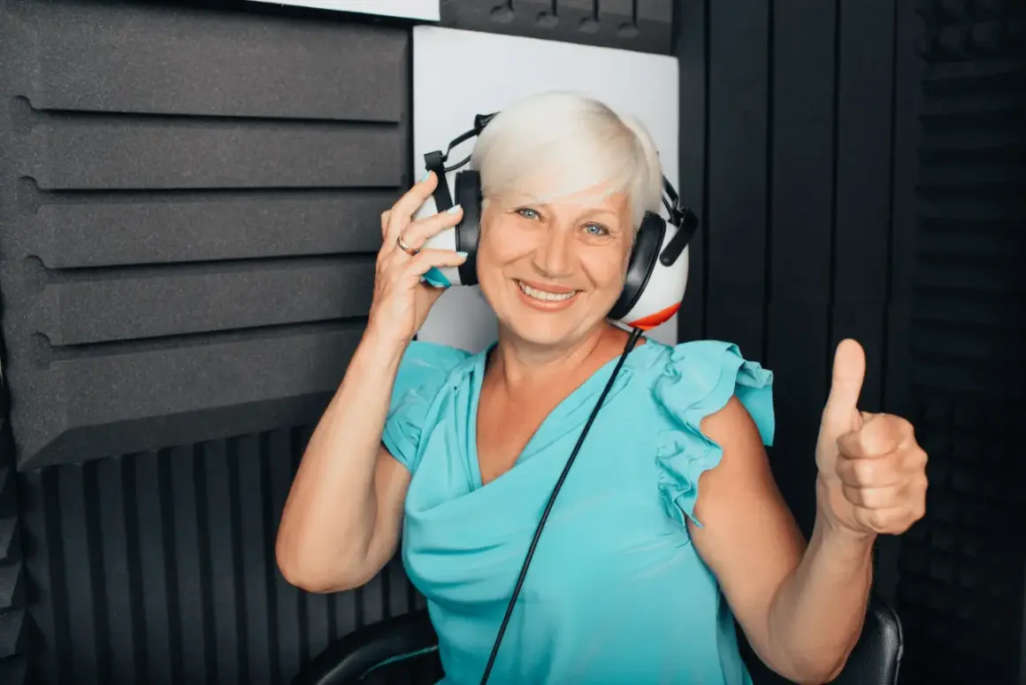 Uśmiechnięta kobieta w słuchawkach podczas badania audiometrii tonalnej. Pokazuje kciuk w górę, co oznacza pozytywny wynik.