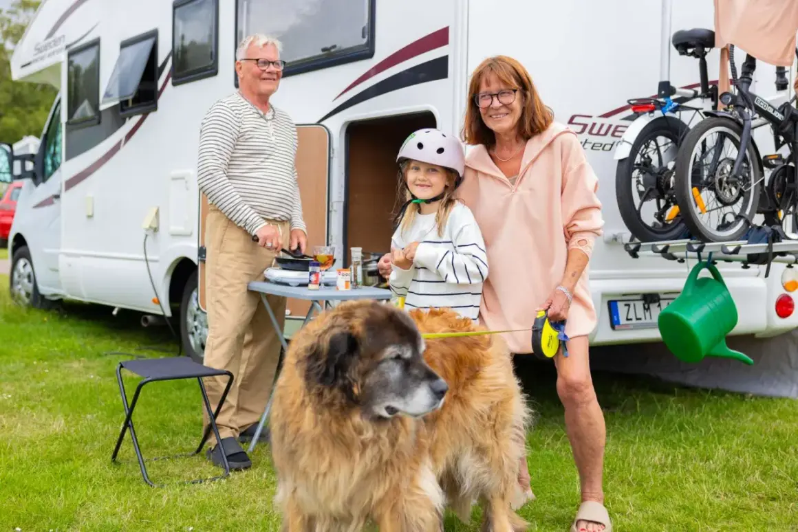 Familjen njuter av att campa med hund vid husbilen. En flicka med hjälm, en man som lagar mat och en kvinna med koppel till hunden.