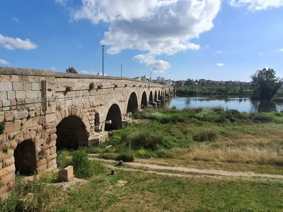 Puente romano de piedra con arcos sobre un río, bordeado de vegetación. Un camino de tierra serpentea por la orilla, evocando la antigua **Vía de la Plata**.