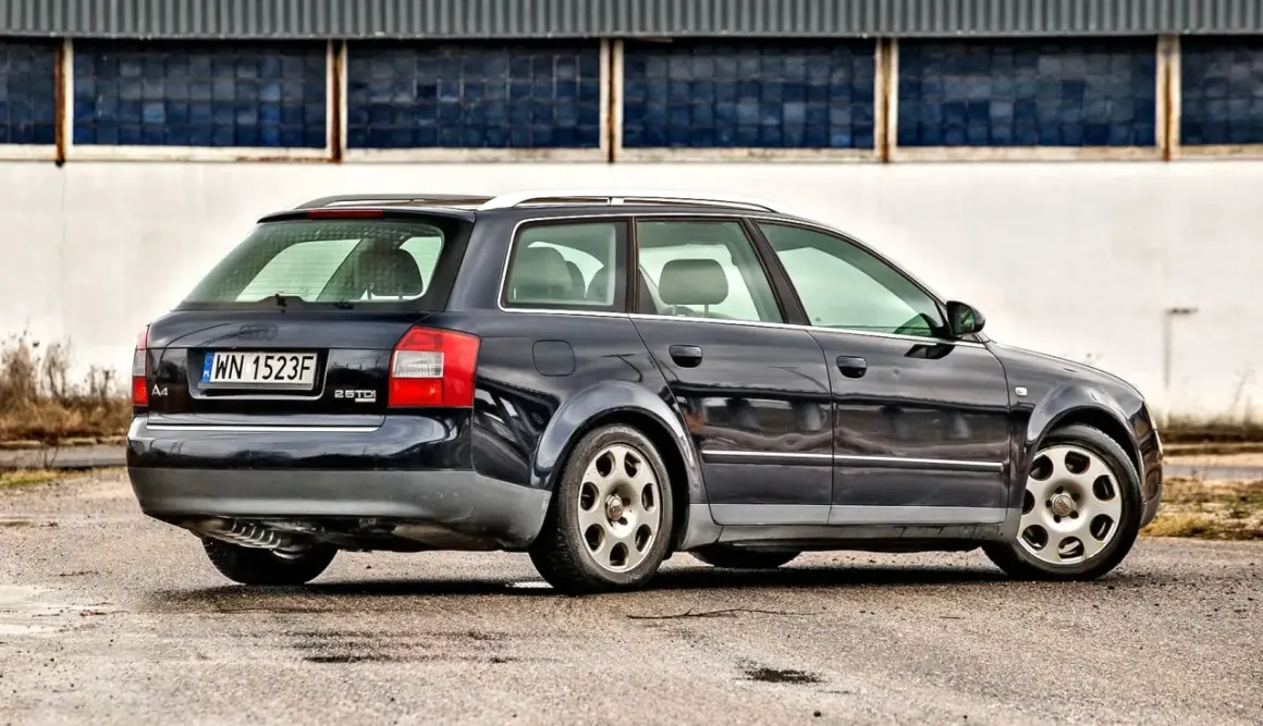 Ciemnogranatowe Audi A4 B6 1.9 TDI Avant stoi na mokrym asfalcie przed budynkiem z niebieskimi oknami.