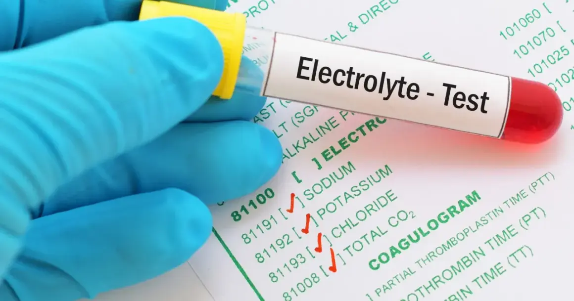 Rękawiczka w niebieskiej rękawiczce trzyma probówkę z napisem "Electrolyte - Test". Obok widać formularz z badaniami, w tym "COAGULOGRAM".
