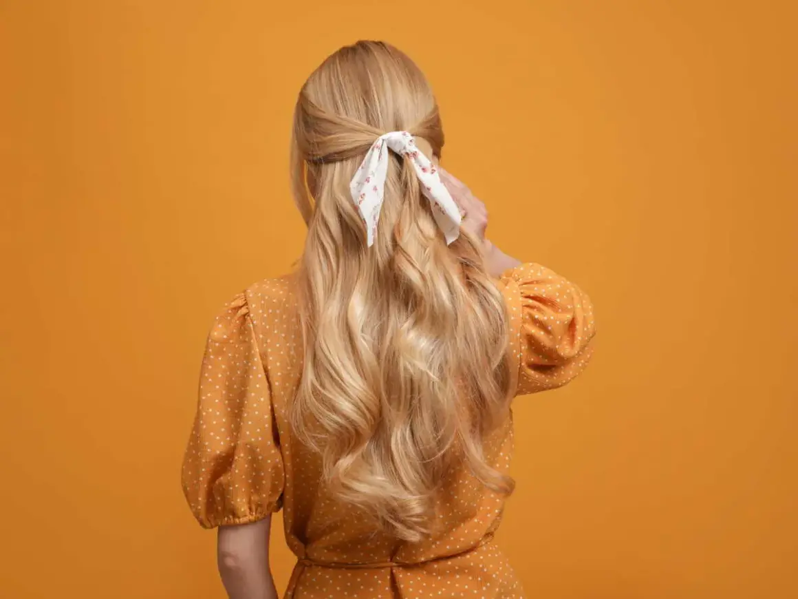 Chevelure blonde ondulée, coiffée en demi-queue avec un foulard fleuri, sur fond orange.