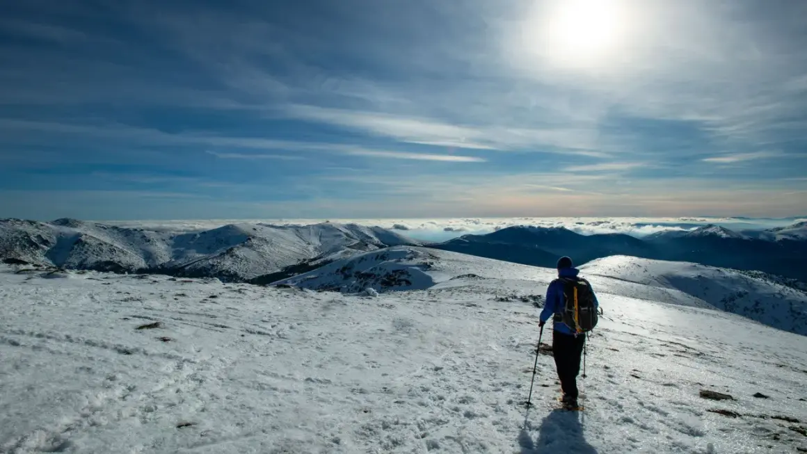 Senderismo en la nieve de Peñalara. Un excursionista con mochila y bastones contempla un paisaje montañoso cubierto de nieve bajo un cielo azul.