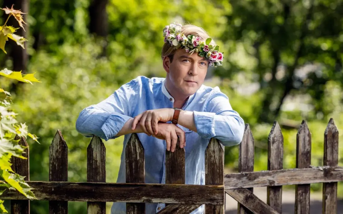 En man med blomsterkrans på huvudet lutar sig mot ett staket. En sommarprat recension som fångar essensen av svensk sommar.