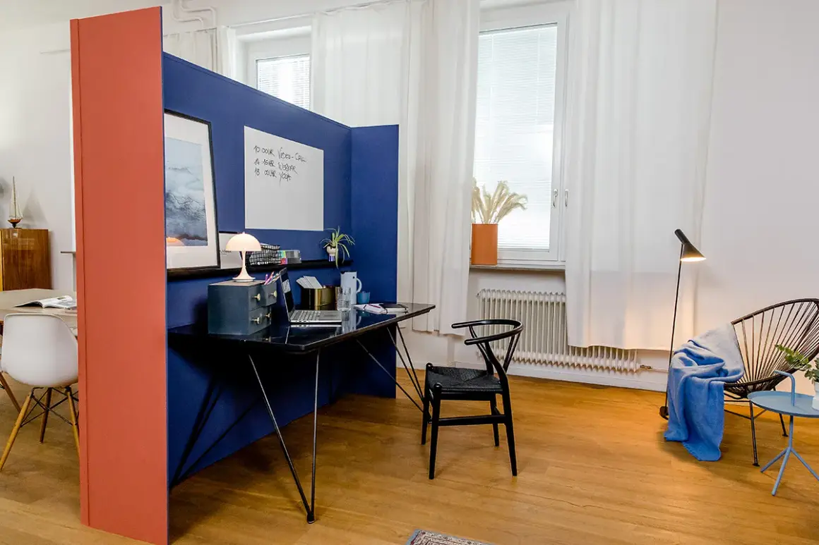 Ein blauer Raumteiler trennt den Arbeitsplatz im Wohnzimmer ab. Ein Schreibtisch mit Laptop und Lampe steht bereit.