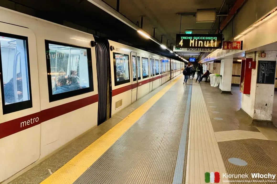 Pasażerowie czekają na peronie na metro we Włoszech. Na tablicy informacyjnej widnieje napis "Treno Successivo 4 Minuti".