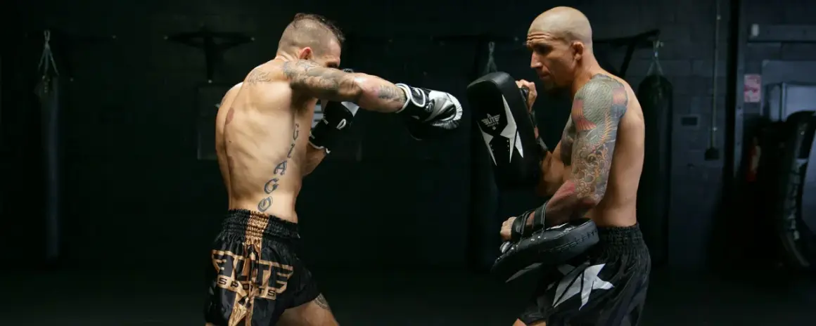 Two men practice boxing without sparring. One throws a punch while the other holds a pad to catch it.