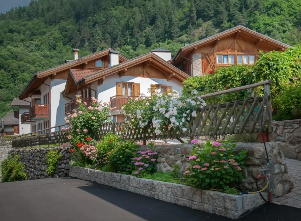 Case con balconi in legno e fiori colorati, incorniciate da un giardino rigoglioso. Un angolo di pace nel Val di Sole, dove si trova questo incantevole paesaggio.