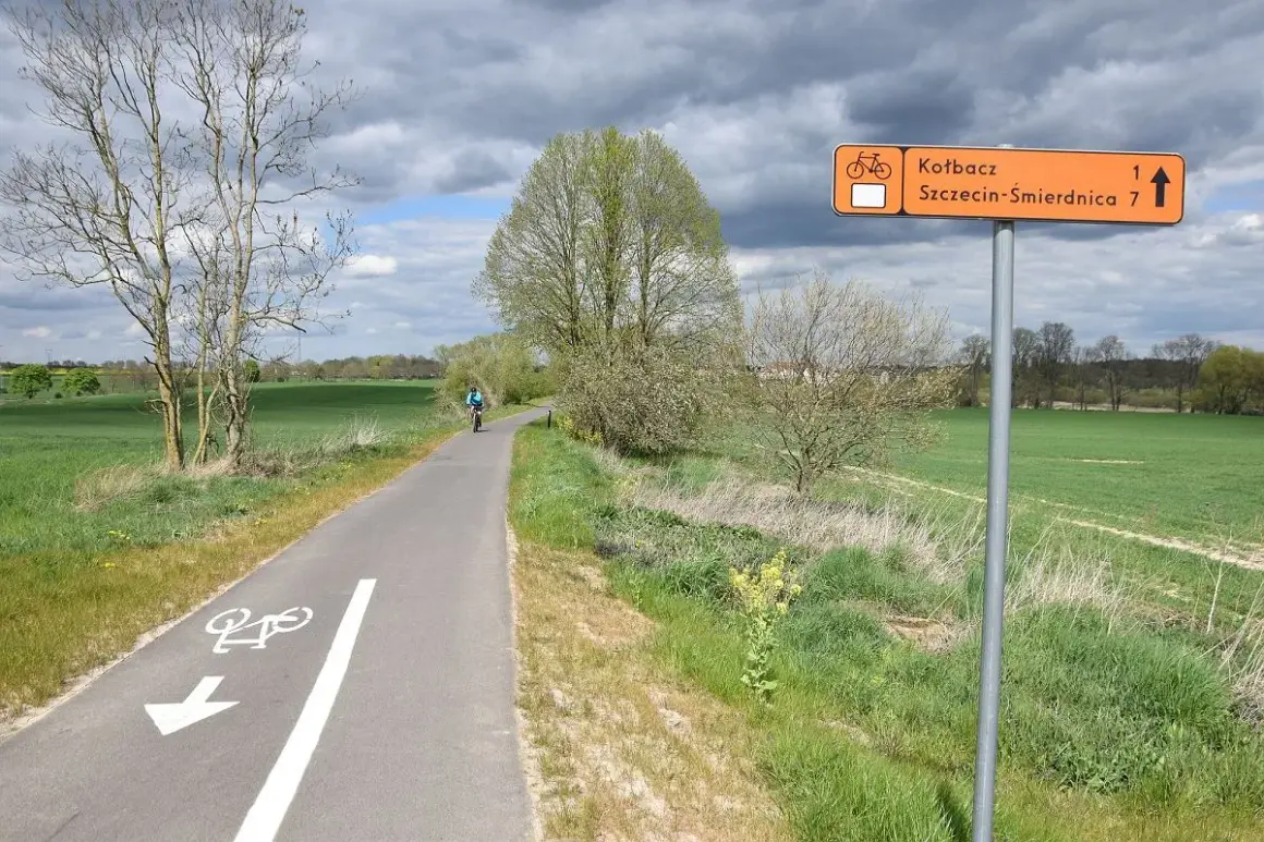 Rowerzysta jedzie ścieżką rowerową przez zielone pola. Pomarańczowy znak wskazuje kierunek do Kotbacza i Szczecin-Śmirdnica.