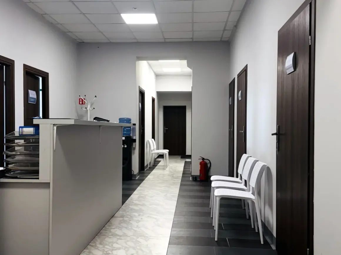 Korytarz kliniki medycznej z recepcją, drzwiami i białymi krzesłami.