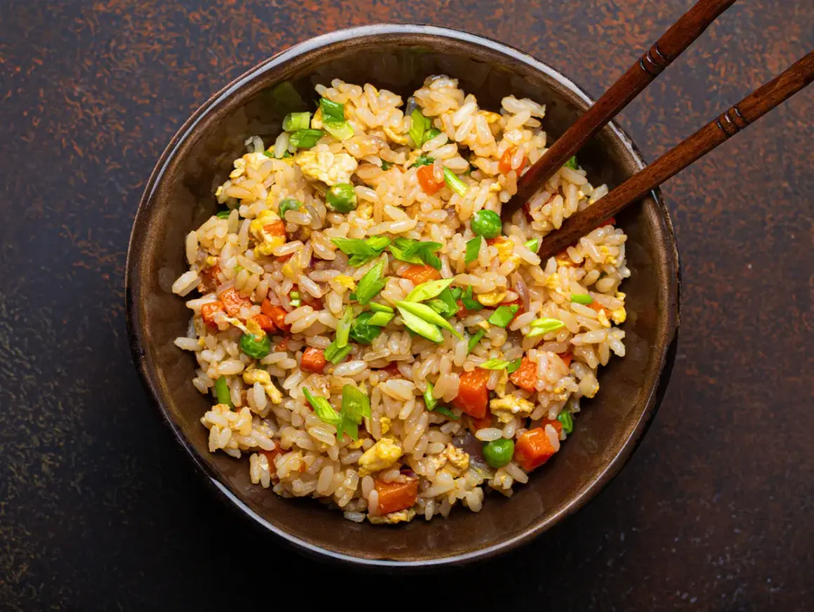 Un piatto fumante di riso alla cantonese ricetta originale, con piselli, carote, uova e cipollotto fresco, pronto per essere gustato con le bacchette.