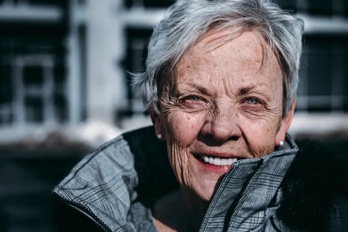 En kvinna med korta frisyrer, dam 65, ler varmt. Hennes grå hår ramar in ett ansikte med vänliga rynkor och blå ögon.