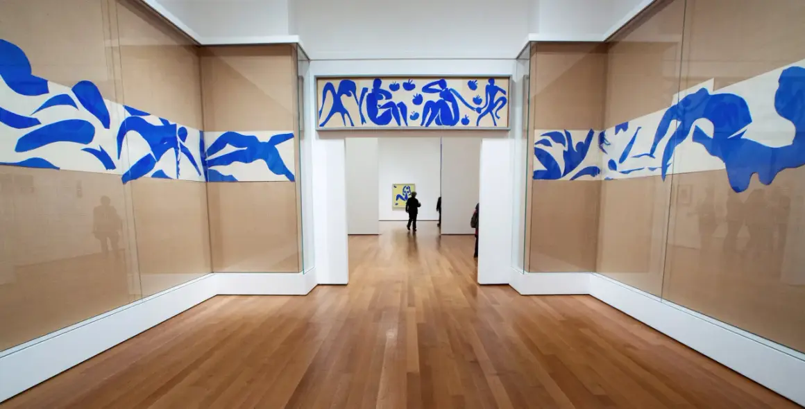 Gallery hallway showcasing Henri Matisse obras: blue cut-outs of figures adorn the walls, with a long horizontal piece above an archway and flowing designs on either side.