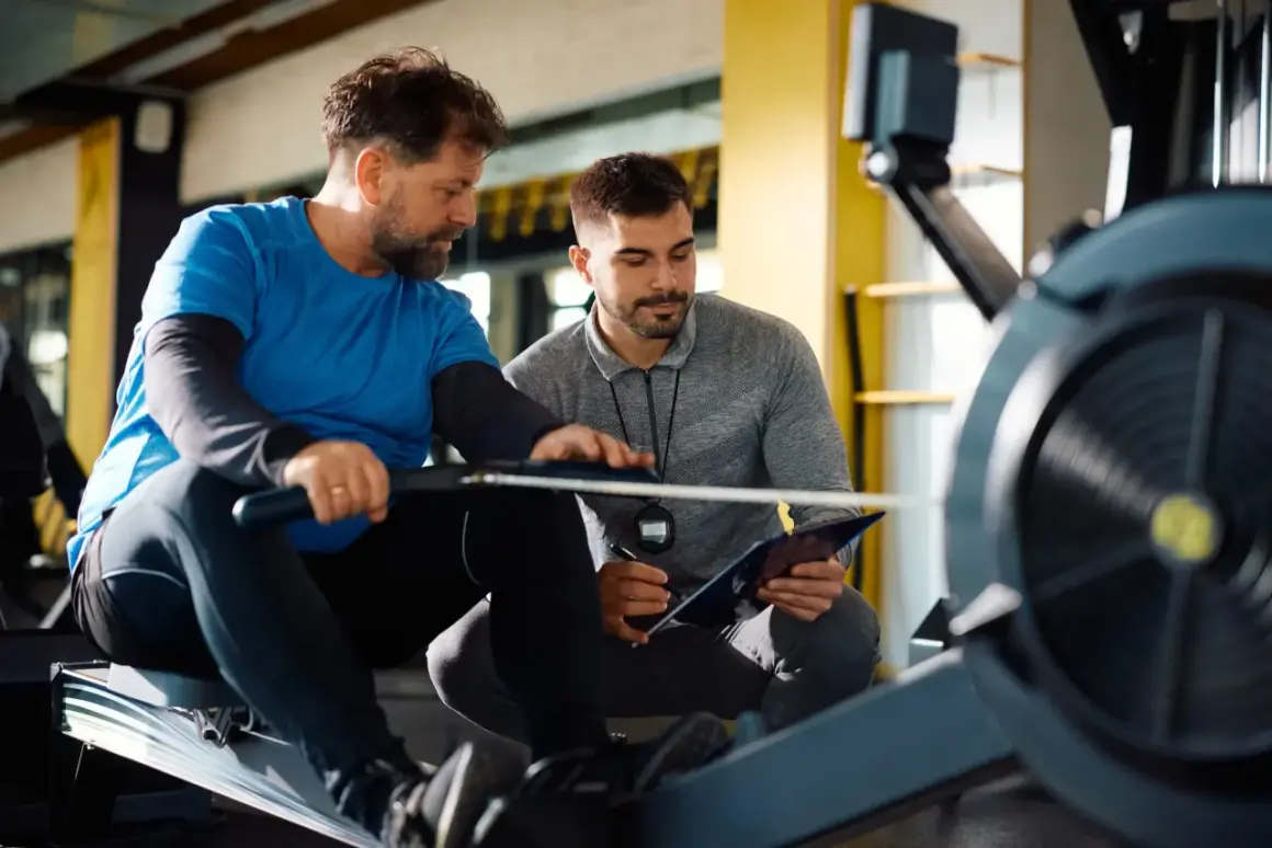 Trener crossfit omawia plan treningowy z podopiecznym na wioślarzu. Zastanawiasz się, ile zarabia trener crossfit?