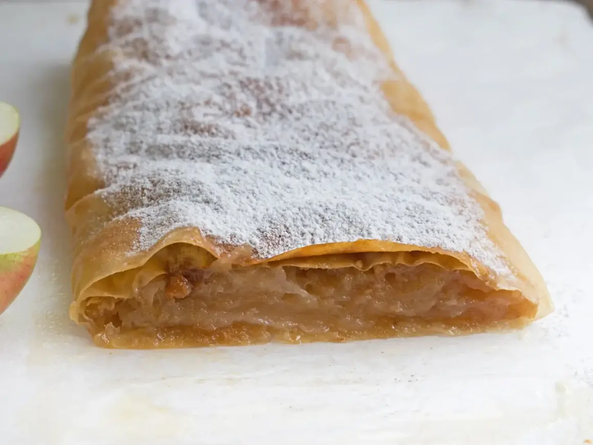 Ein köstlicher Apfelstrudel mit fertigem Strudelteig, bestäubt mit Puderzucker. Daneben liegen frische Apfelstücke.