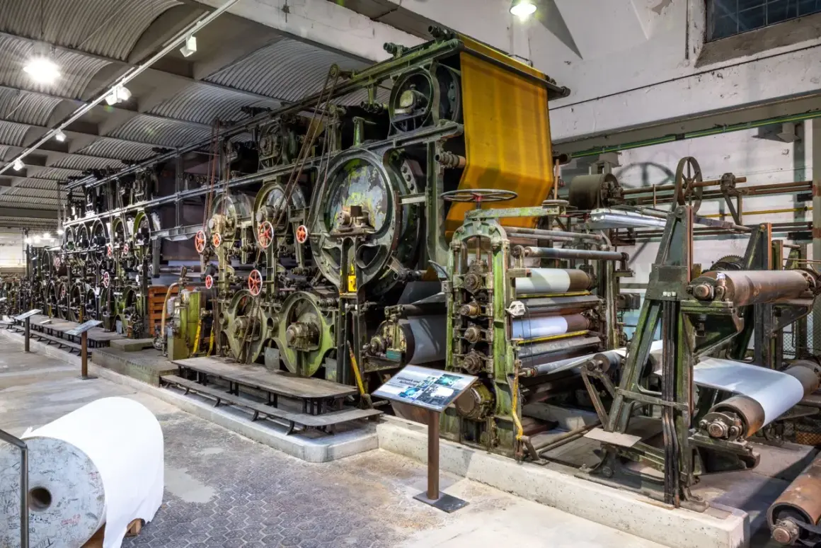 Große, grüne Textilmaschine im lvr industriemuseum, die Stoffbahnen verarbeitet. Eine gelbe Stoffbahn hängt oben, daneben Papierrollen.