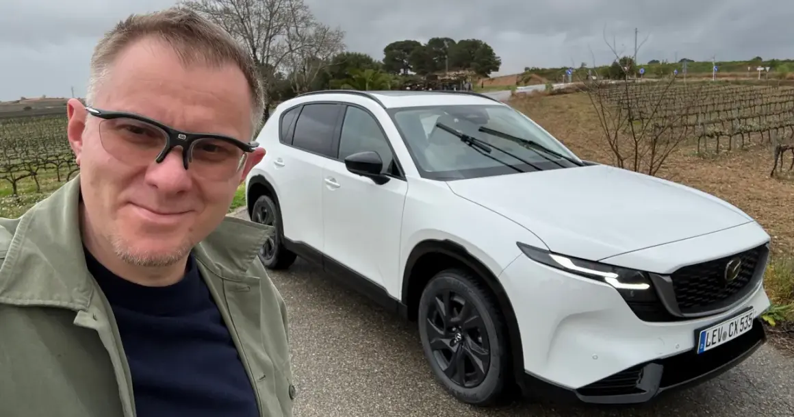 Mężczyzna w okularach pozuje do selfie obok białej Mazdy CX-5 na tle winnicy.
