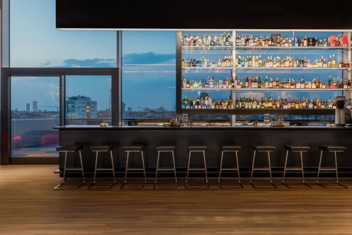 Interni di un locale con bancone bar, sgabelli e scaffali pieni di bottiglie. Vista sulla città al tramonto, un'atmosfera da next club Torino.