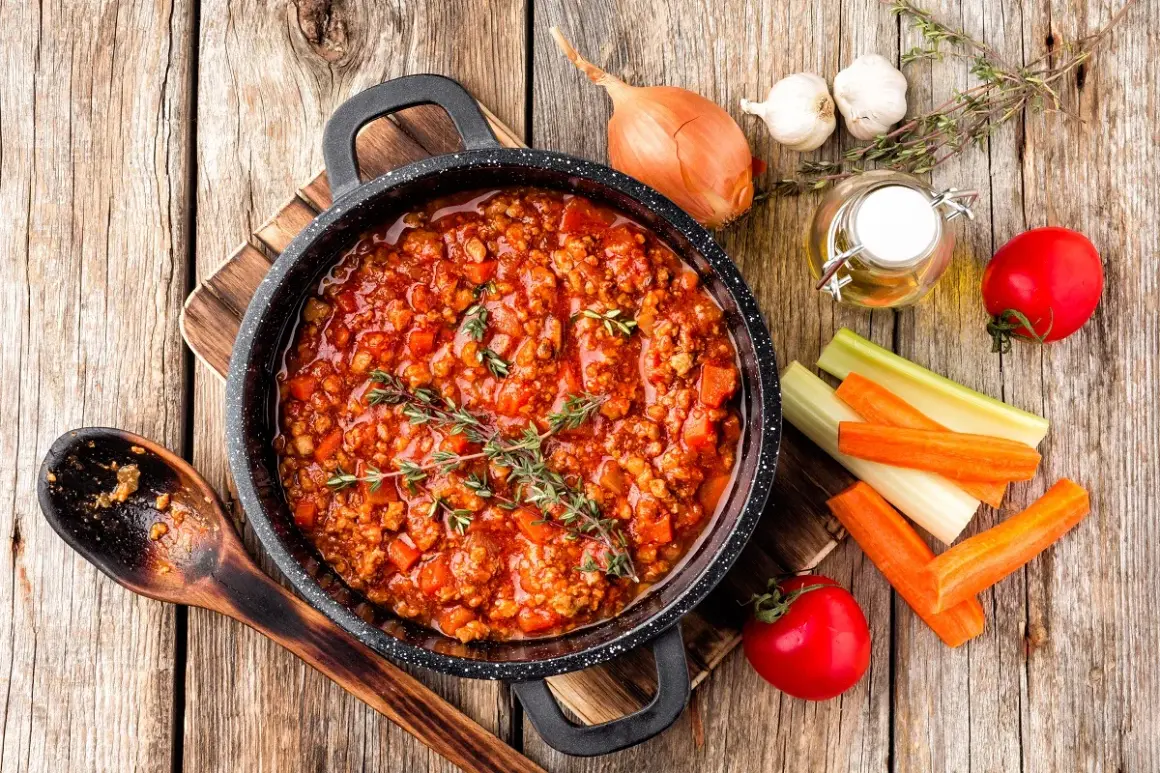 Pyszny sos bolognese w garnku, gotowy do podania z makaronem. Obok składniki: cebula, czosnek, marchew, seler, pomidory i oliwa. Idealny pomysł, co zrobić z sosem bolognese!