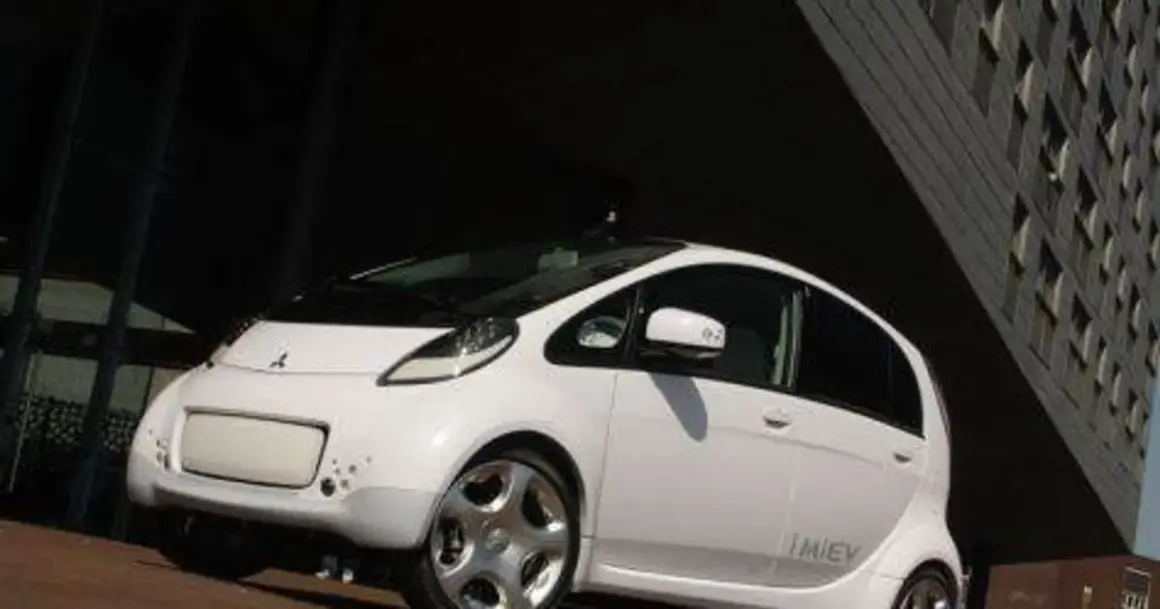 Biały, kompaktowy Mitsubishi i-MiEV stoi przed nowoczesnym budynkiem.