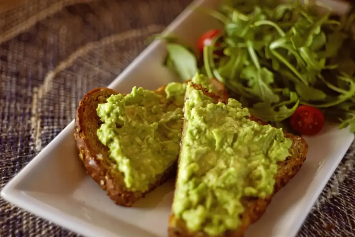 Dwie kromki pełnoziarnistego chleba z najpyszniejszym guacamole, obok sałatka z rukoli i pomidorki koktajlowe.