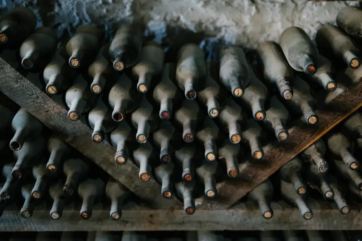 Des bouteilles de vin poussiéreuses, empilées sur des étagères en bois. La cave à vin semble avoir un problème de température, les bouchons sont secs.