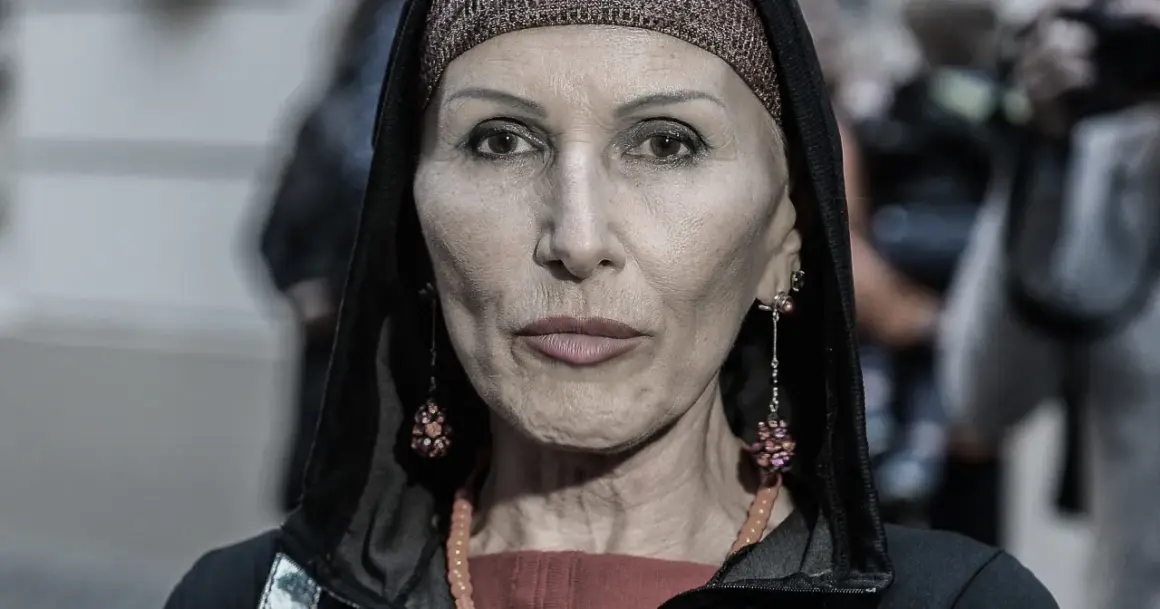 Kobieta w kapturze i ozdobnej opasce na głowie. Jej twarz, naznaczona kora wiek, opowiada historię. Długie kolczyki i naszyjnik dodają jej charakteru.