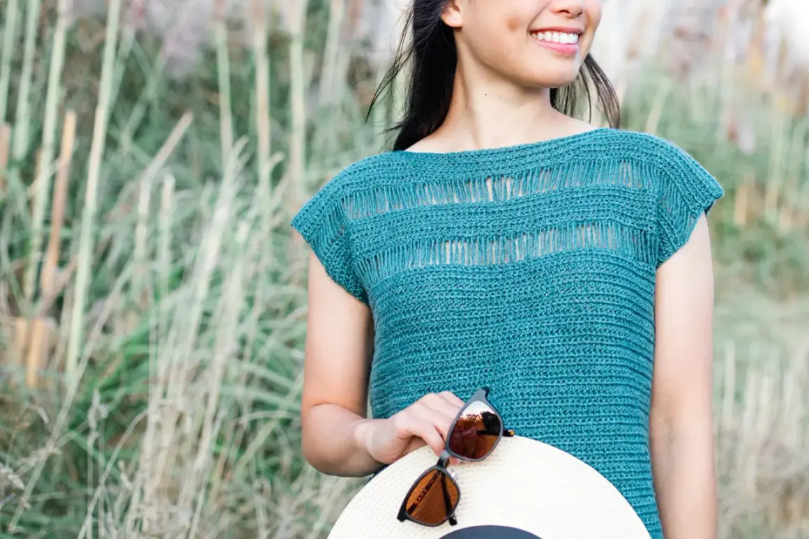 Donna sorridente con un top uncinetto blu petrolio, tiene un cappello di paglia e occhiali da sole.