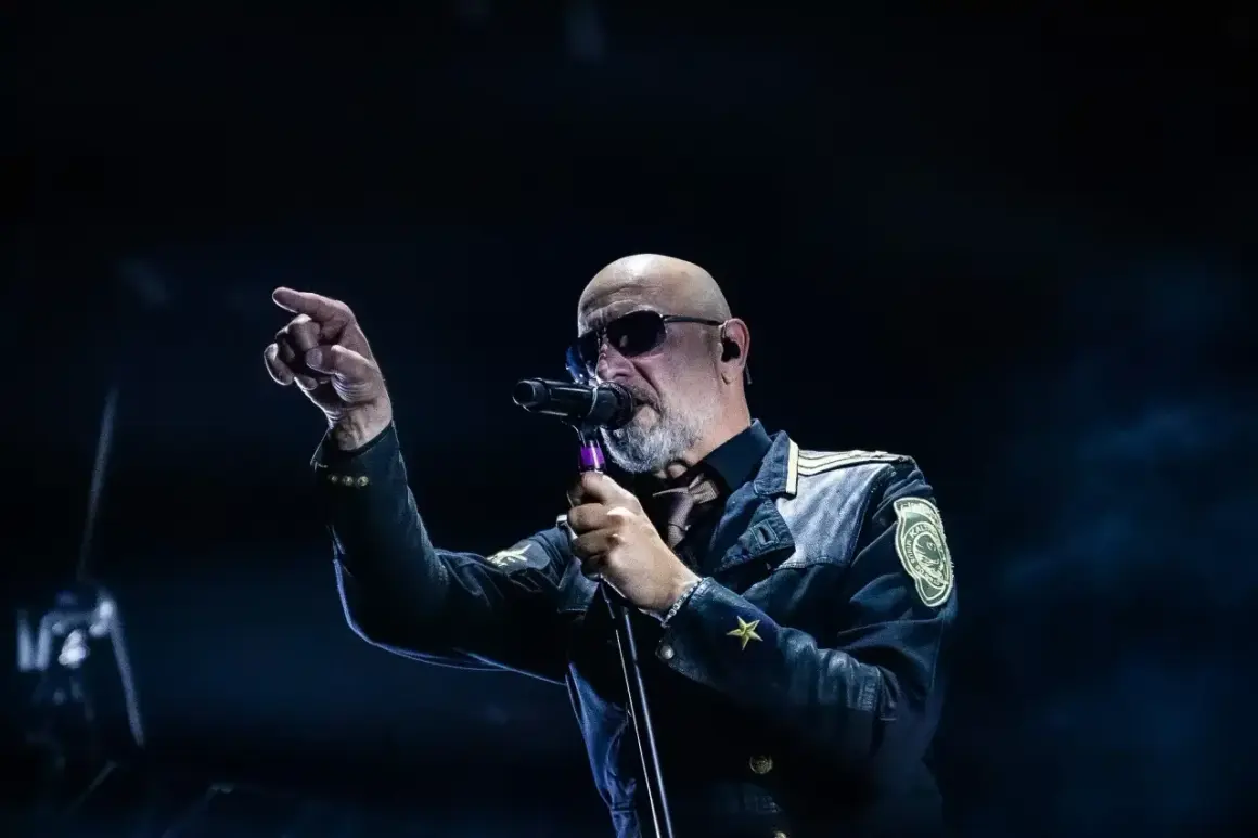 Der Sänger von Eisbrecher Ludwigsburg, mit Sonnenbrille und markanter Jacke, singt ins Mikrofon und zeigt mit dem Finger ins Publikum.