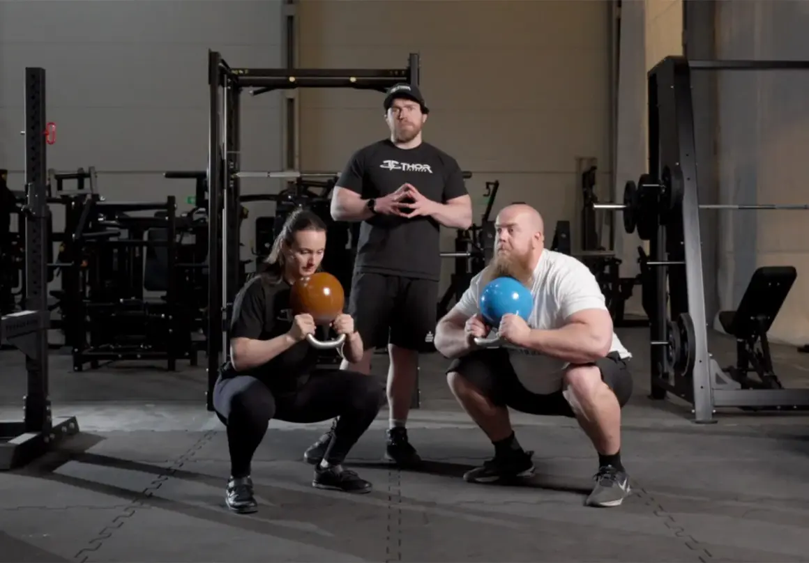 Två personer utför goblet squat med kettlebells medan en instruktör står bakom.