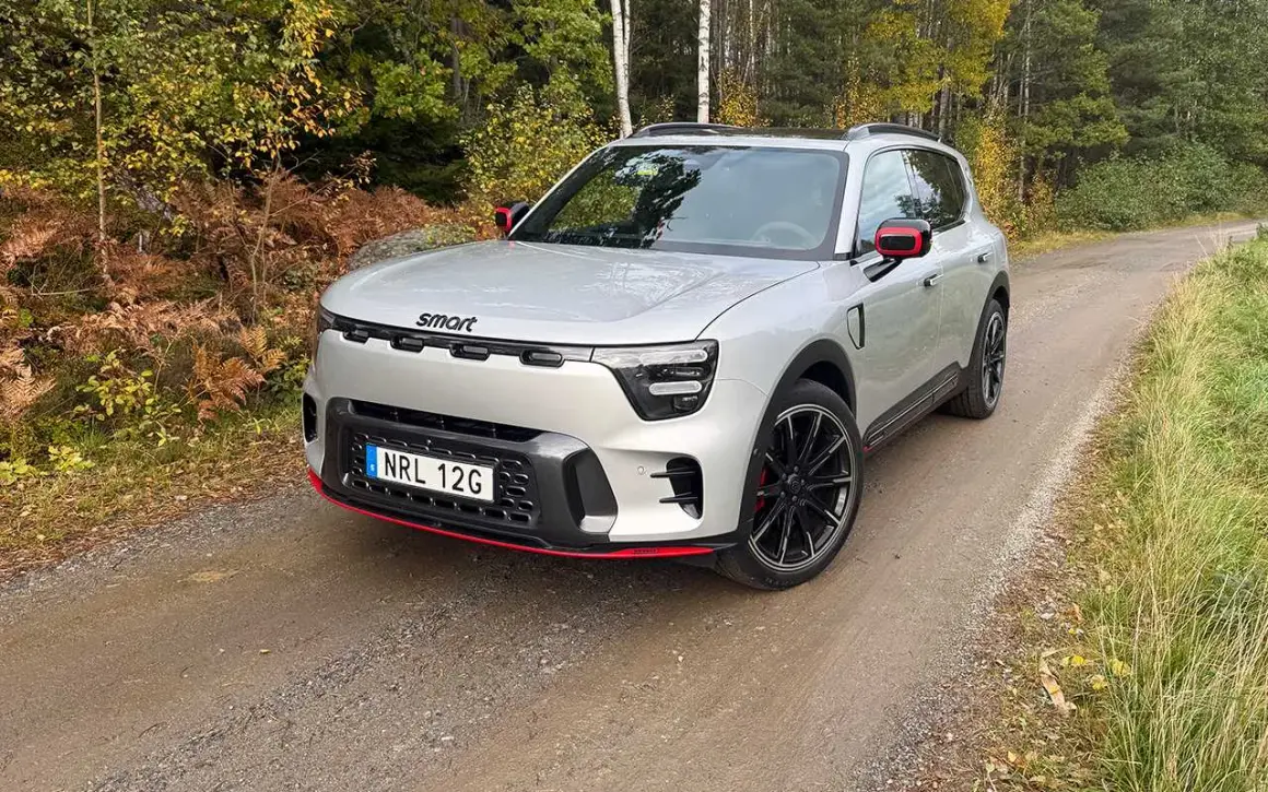 En smart 5-dörrars SUV i silver med röda detaljer kör på en grusväg genom en höstskog.