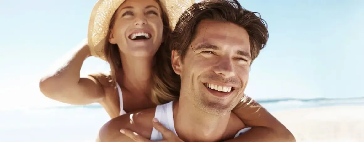 Glückliches Paar am Strand, die Frau lacht und trägt einen Hut. Selbst im Schatten wird man braun, wenn man die Sonne genießt.