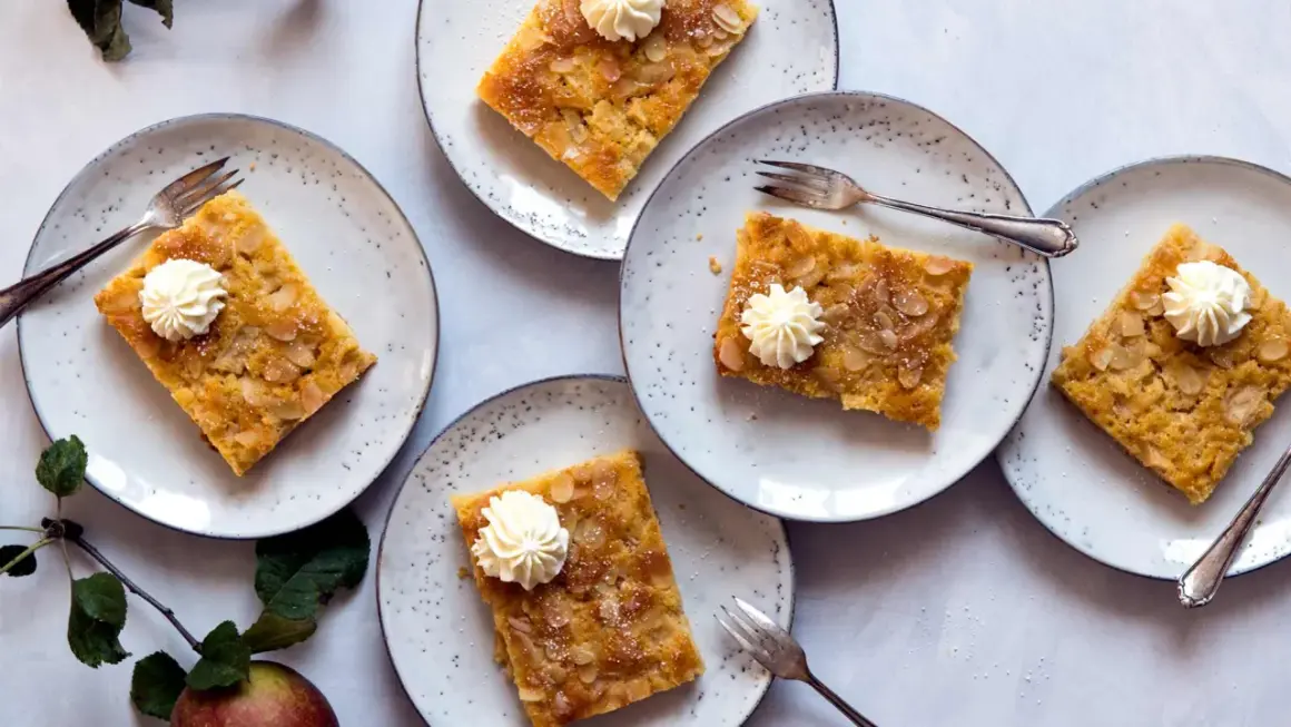 Saftiger Apfelkuchen vom Blech, garniert mit Sahnetuffs und Mandelblättchen, auf Tellern angerichtet.