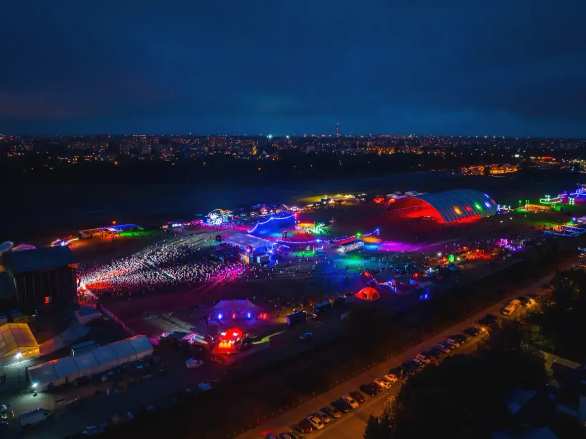 Nocne, kolorowe światła rozświetlają teren festiwalu na powietrzu. Tłumy bawią się przy scenach i namiotach, a w tle widać miasto.