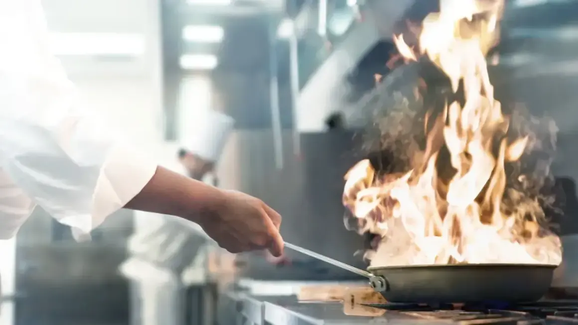 Ein Koch wirbelt eine brennende Pfanne in einer professionellen Küche. Flammen schlagen hoch und erzeugen viel Rauch.