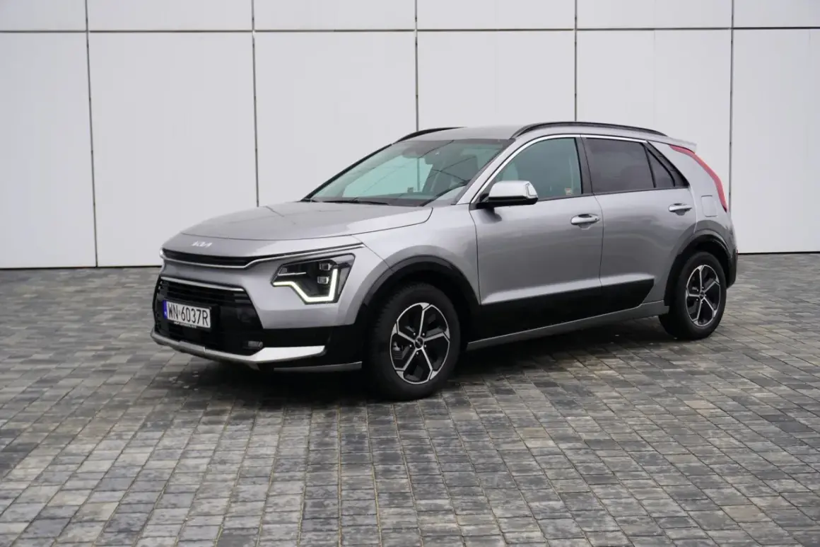 Srebrny Kia Niro na tle białej ściany. Wiele osób szuka kia niro opinie przed zakupem.