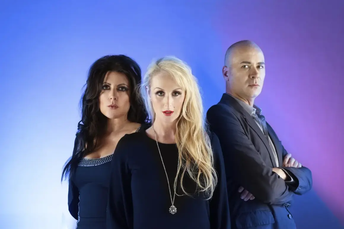 Miembros de The Human League: dos mujeres con maquillaje dramático y un hombre calvo con traje, posando ante un fondo azul y morado.