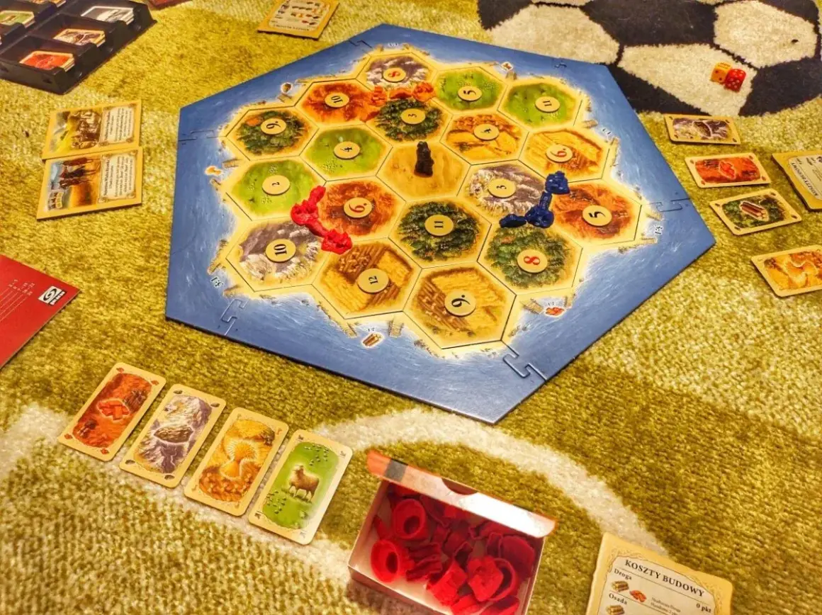 Plansza Catan: Świt Ludzkości, karty surowców i pionki. Rozgrywka w toku, gracze budują osady i drogi.