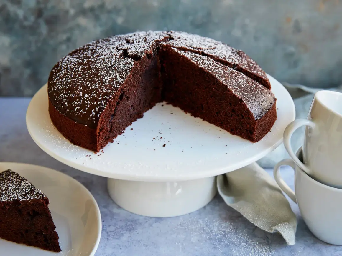 Ein saftiger veganer Schokokuchen, bestäubt mit Puderzucker, steht auf einem weißen Tortenständer. Daneben liegen Tassen und eine Serviette.