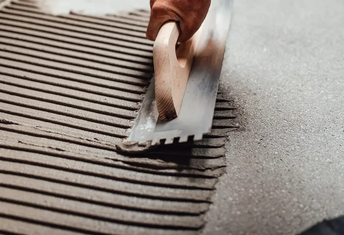 Application d'enduit pour carrelage collé avec une truelle crantée. Les sillons réguliers préparent la surface pour une pose parfaite.