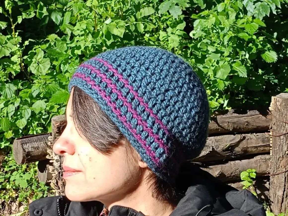 Donna con cappello uncinetto tutorial, blu e fucsia, con sfondo verde e tronchi.