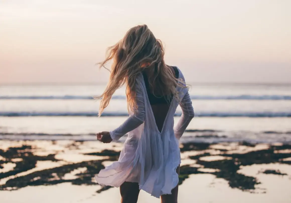 Blondynka w zwiewnej białej sukience tańczy na plaży, a wiatr rozwiewa jej włosy. W tle widać spokojne beach waves i zachodzące słońce.
