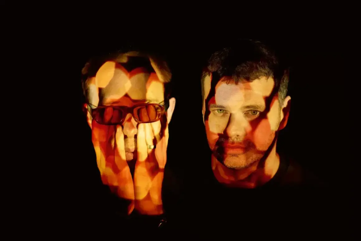 The Chemical Brothers logo is projected onto the faces of two men, one wearing sunglasses, in a dark, atmospheric setting.